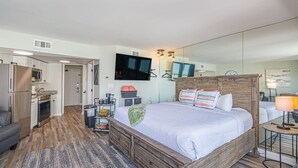 Un dormitorio con cama, televisor, espejo, escritorio y kitchenette.