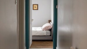 En smal gang der fører til et soveværelse med en seng, et enkelt billed ophængt på væggen, og en dør med turkis kant.