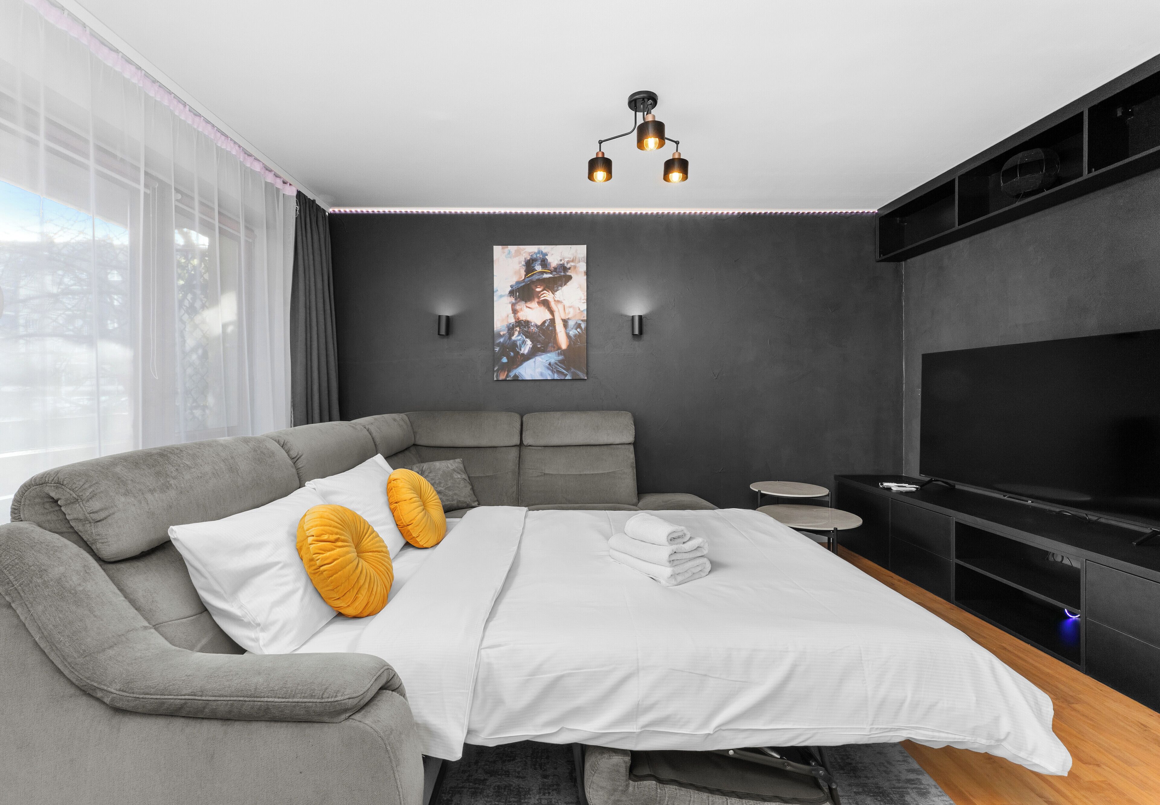 Een moderne slaapkamer met een grijze bank, een bed met witte beddengoed, een schilderij aan de muur en een televisie aan de rechterkant.