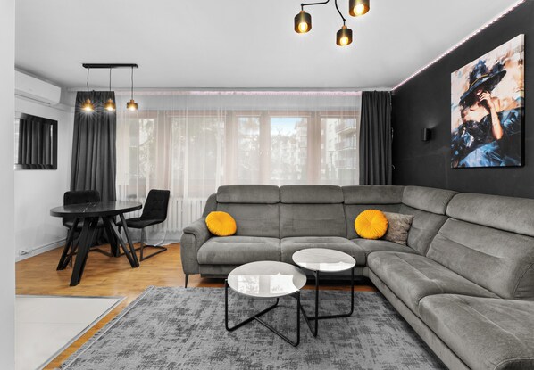A modern living room with a grey sectional sofa, two round tables, a dining table with chairs, and a large window with sheer curtains.
