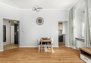 Une pièce spacieuse avec un plancher en bois, une table à manger avec des chaises, une horloge murale et un coin cuisine visible à travers une porte ouverte.