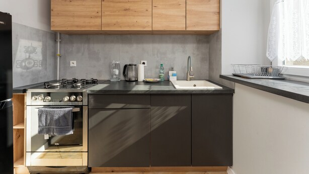 A kitchen with a built-in oven, a sink, wooden cabinets, and a window with lace curtains. - Daszyńskiego Urban Comfort Apartment (Gliwice)