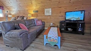 Een kamer met houten lambrisering, een bank, een televisie en een tafeltje met een tafelkleed in oogstthema.