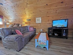 A wooden-paneled room with a sofa, a television, and a small table with a harvest-themed tablecloth. - Hideaway Cabin Bryson City, NC - 5 Minutes from Downtown (Bryson City)