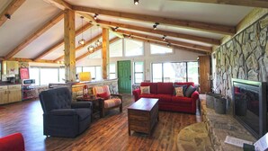 A spacious living area with a fireplace, wooden beams, a stone wall, and a kitchen in the background. - View 280- Amazing views of the Smoky Mountains (Bryson City)