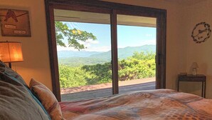 A bedroom with a large bed, a nightstand, a lamp, and a view of a mountainous landscape through a sliding door. - View 280- Amazing views of the Smoky Mountains (Bryson City)