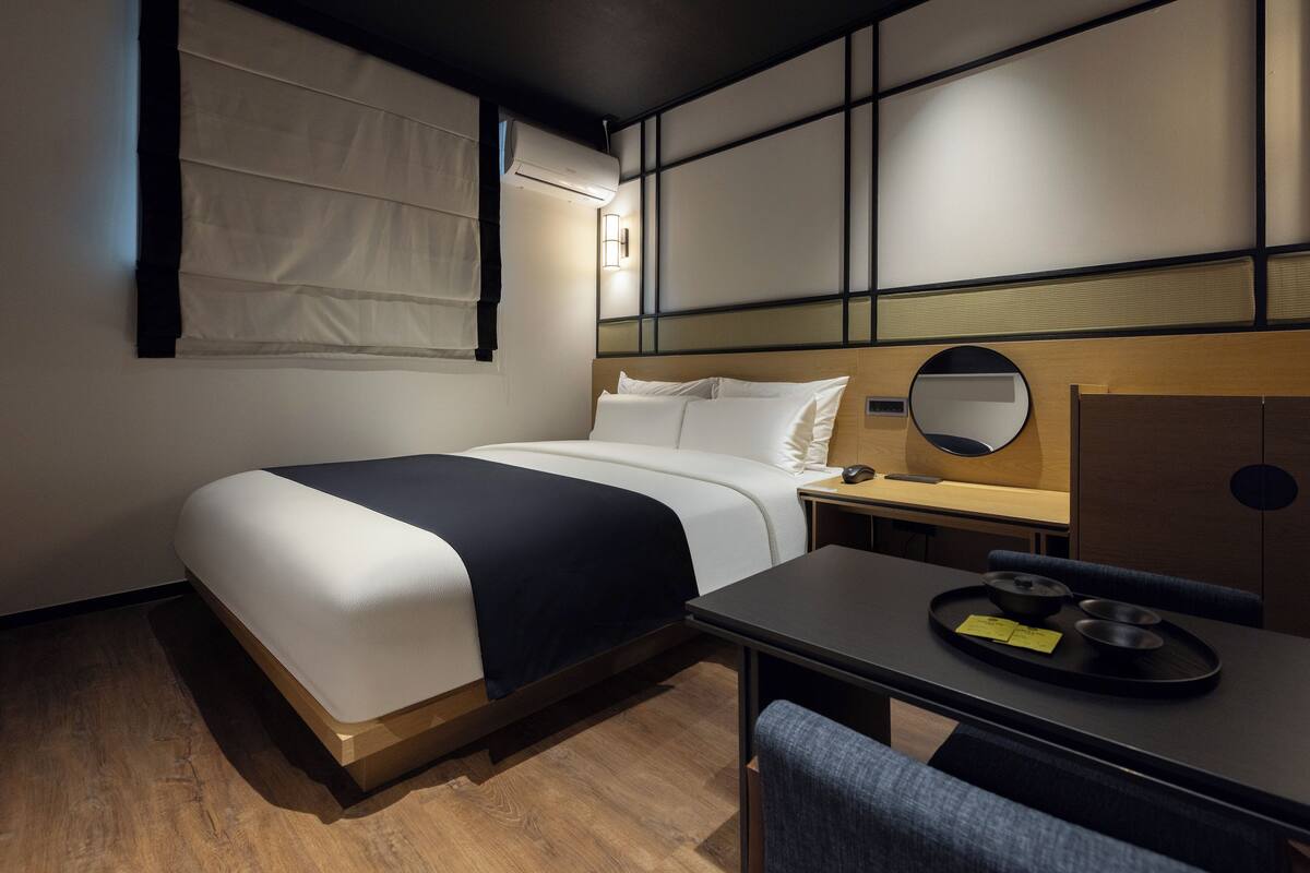 Quarto de hotel com cama, mesa de cabeceira, cadeira e escrivaninha com bandeja.