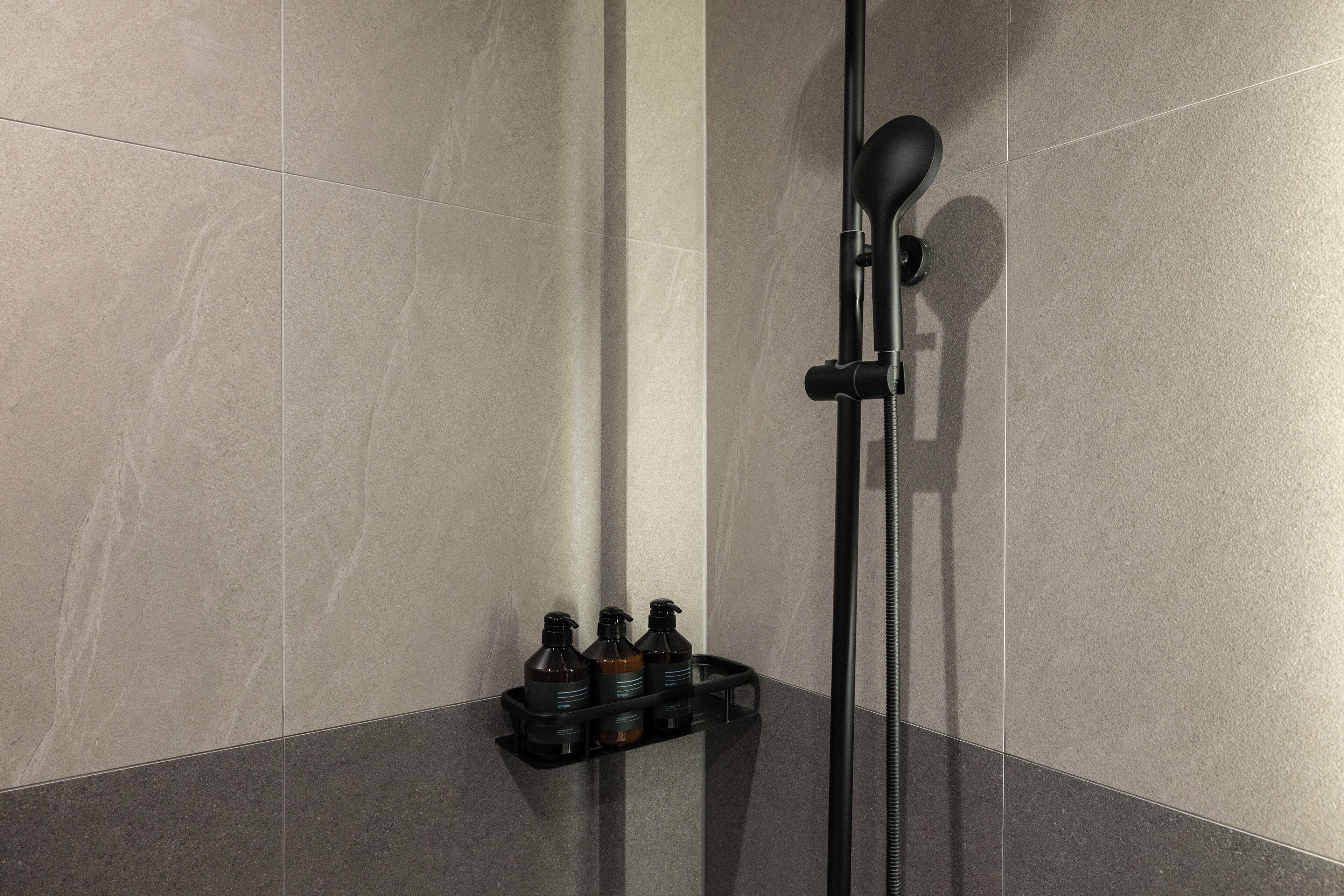 Un espace douche équipé d’un pommeau de douche noir à main, d’un tuyau d’eau vertical et d’une étagère contenant trois bouteilles.