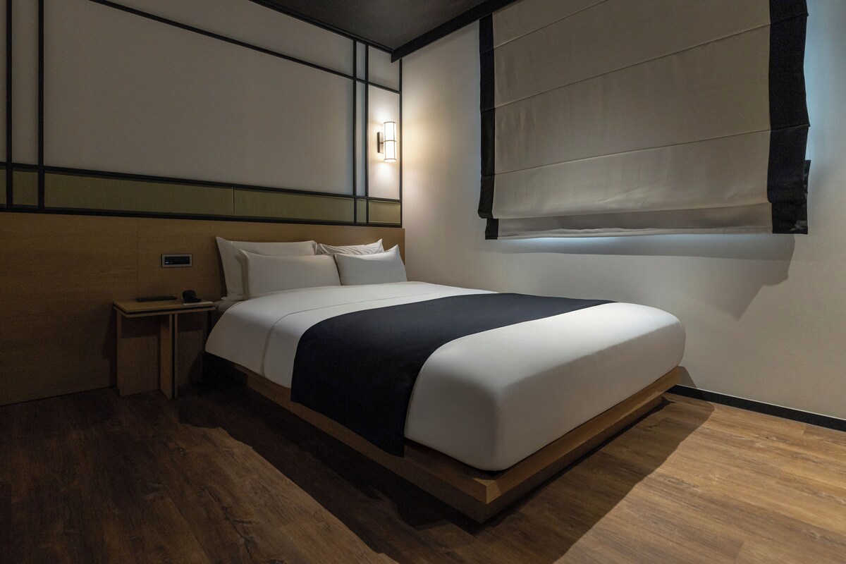 A hotel room with a large bed, bedside tables, a wall-mounted light, and a window with blinds.