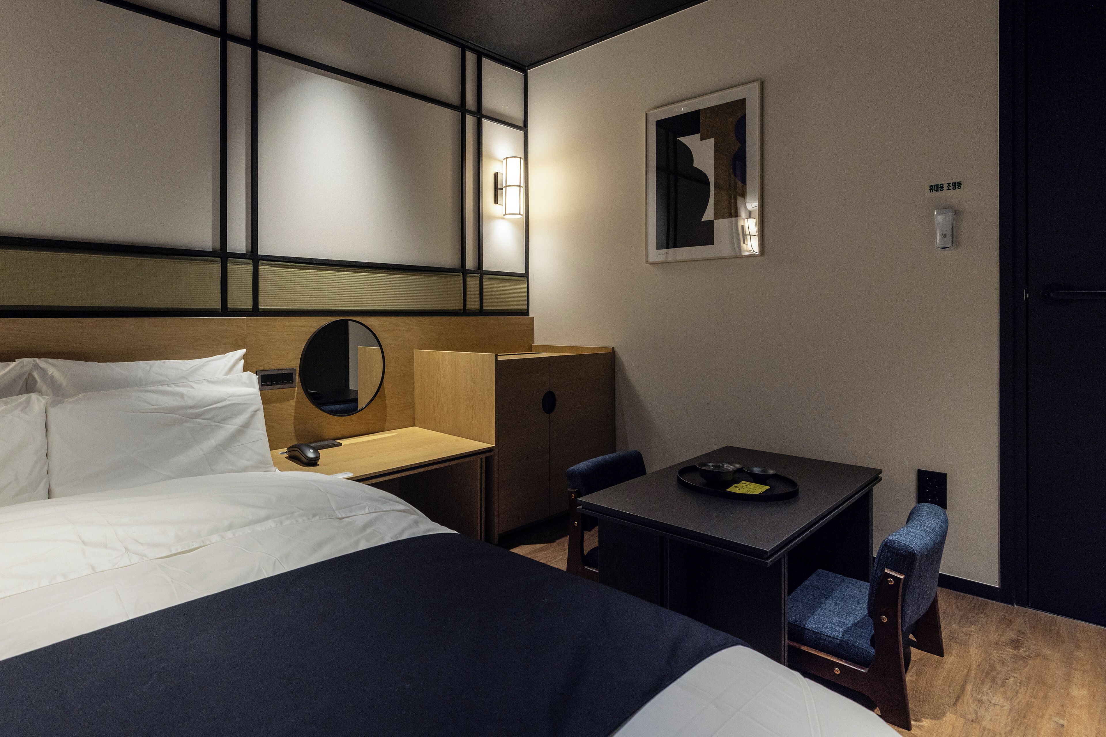 A hotel room with a bed, a desk, a chair, a mirror, and a wall-mounted light fixture.