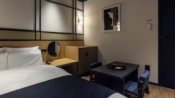 A hotel room with a bed, a desk, a chair, a mirror, and a wall-mounted light fixture.