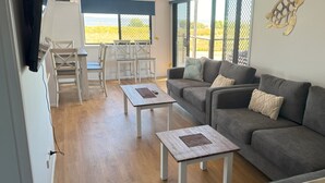 A living room with a grey sofa, two white tables, and a dining area with chairs. - 3 Bedroom, 2 Bathroom Holiday House (Karumba)