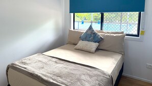 A bedroom with a bed, a window with a blue blind, and a view of a pool outside. - 3 Bedroom, 2 Bathroom Holiday House (Karumba)