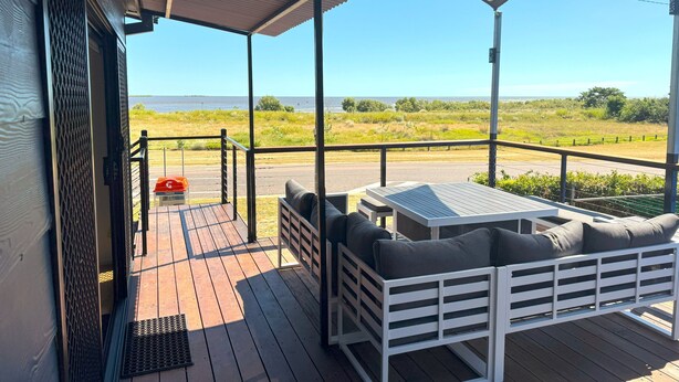 A covered outdoor area with a wooden deck, a white bench with cushions, and a view of the sea. - 3 Bedroom, 2 Bathroom Holiday House (Karumba)