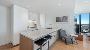 A modern kitchen with a central island, a dining area with a chair and table, and a large window offering a city view. - Skyline Apartment in Fortitude Valley (Fortitude Valley)