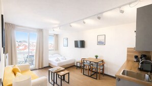 Una habitación de hotel moderna con sofá, una mesa de comedor con sillas, cama, televisor y una zona de cocina con lavamanos.