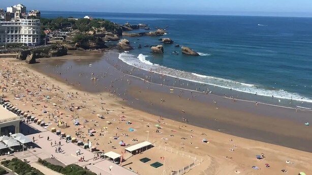 Una playa con muchas personas, cielo despejado y afloramientos rocosos en el agua.