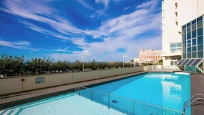Una piscina bajo un cielo azul despejado, un edificio blanco con amplias ventanas y un edificio rojo al fondo.