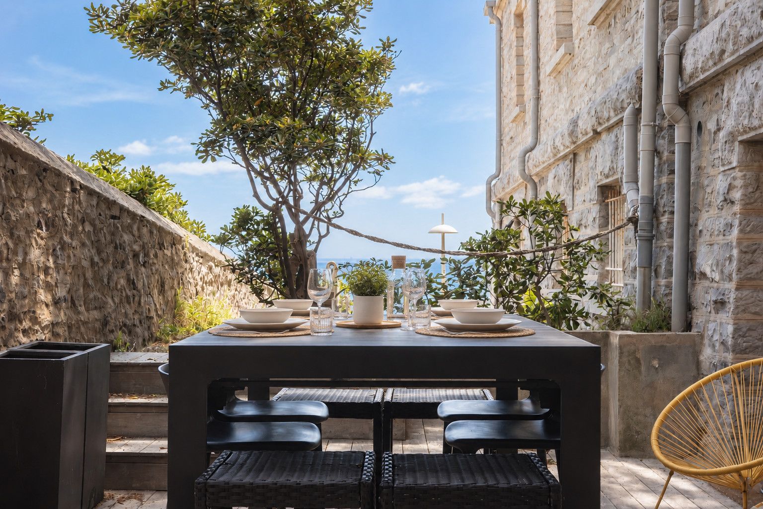 An outdoor dining area with a table set for four, surrounded by stone walls and a view of the sea.