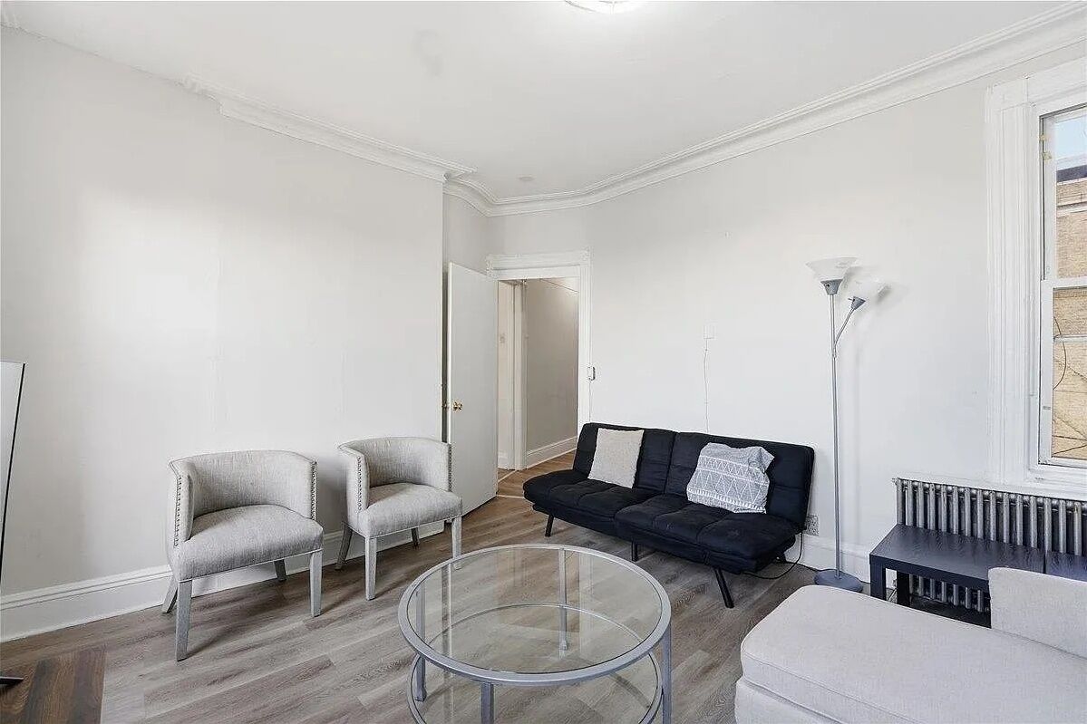 Een woonkamer met een zwarte bank, twee fauteuils, een glazen salontafel en een radiator.