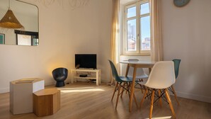 Een moderne woonkamer met een televisie op een wit meubel, een eettafel met stoelen, een ronde spiegel en een groot raam met gordijnen.