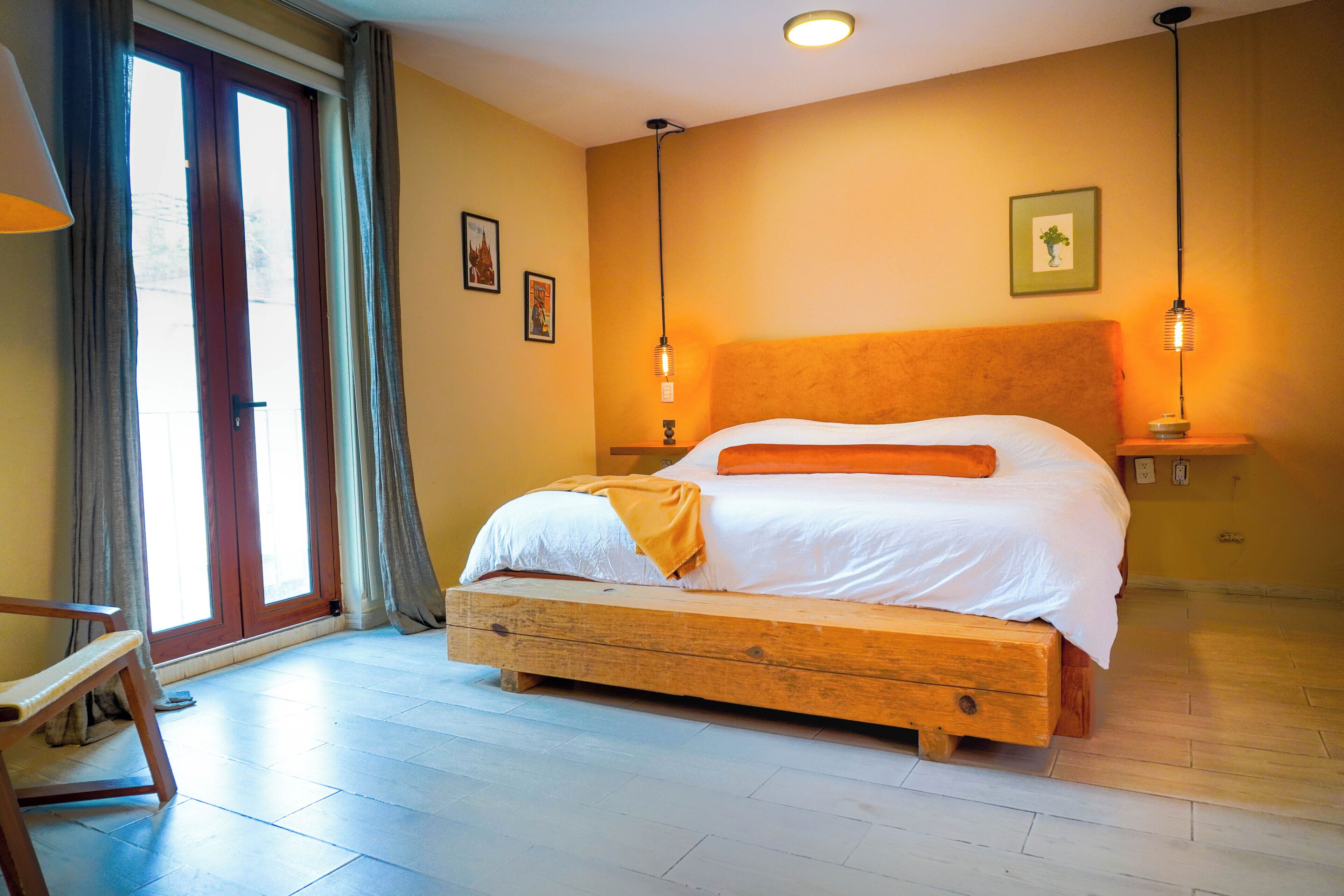 Un dormitorio con cama de madera, un banco, una ventana con cortinas e iluminación en la pared.