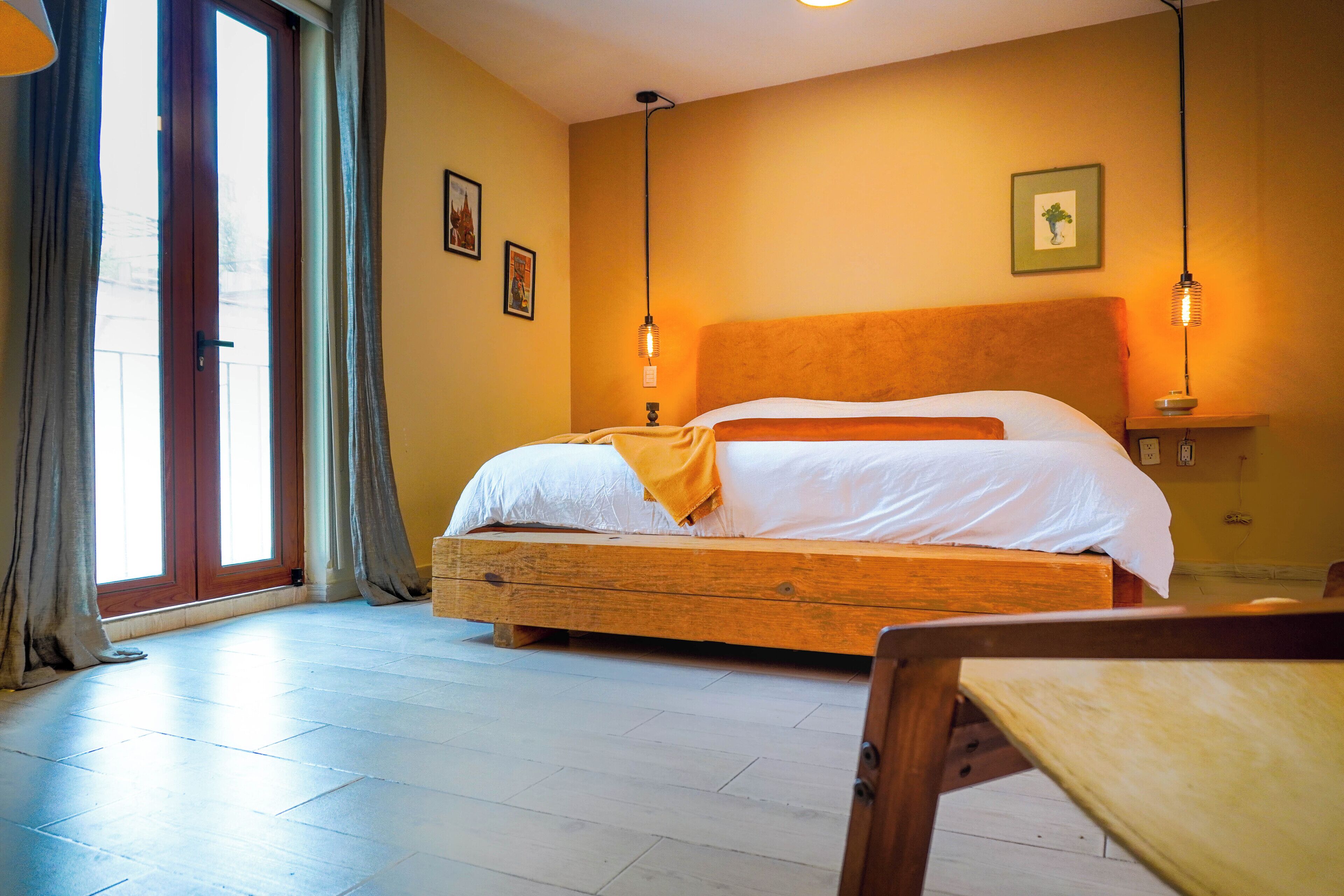 A bedroom with a wooden bed, a large window with curtains, and a bedside table with a lamp.