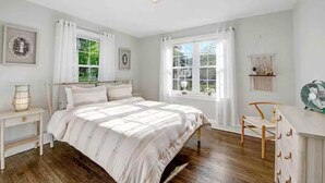 Un dormitorio con una cama, dos ventanas, una mesita de noche, una silla y un ventilador.