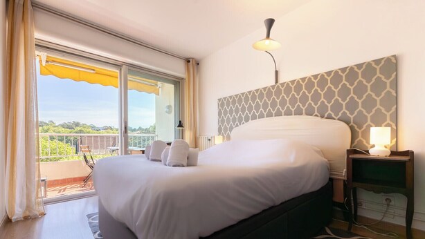 A bedroom with a large bed, a nightstand with a lamp, a view of the outdoors through a sliding door, and a patterned wall. - Cosy apartment with Balcony near the Beach (Biarritz)