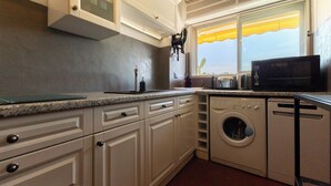 A kitchen with white cabinets, a washing machine, a microwave, and a window with a yellow awning. - Cosy apartment with Balcony near the Beach (Biarritz)