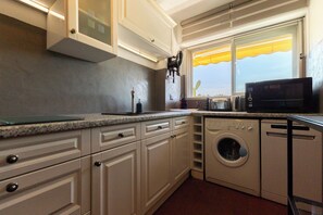 A kitchen with white cabinets, a washing machine, a microwave, and a window with a yellow awning. - Cosy apartment with Balcony near the Beach (Biarritz)