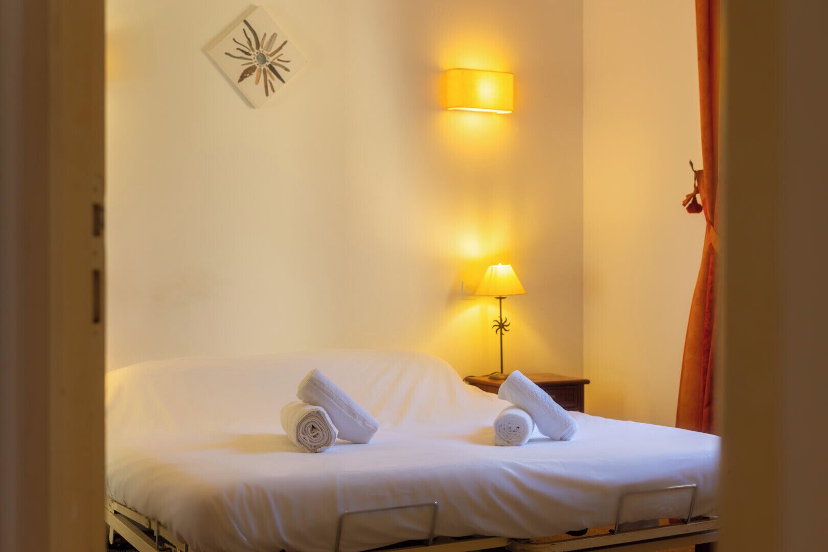 A hotel room with a bed, two rolled towels, a wall-mounted lamp, and a decorative wall piece.