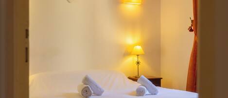 A hotel room with a bed, two rolled towels, a wall-mounted lamp, and a decorative wall piece.