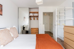 A bedroom with a bed, a dresser, a closet with hanging clothes, and a door leading to another room. - Blueground | Poplar, balcony, nr Thames (London)