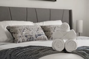 Un lit bien fait, avec des serviettes blanches pliées en rouleau, des coussins gris à motifs et une couverture grise.