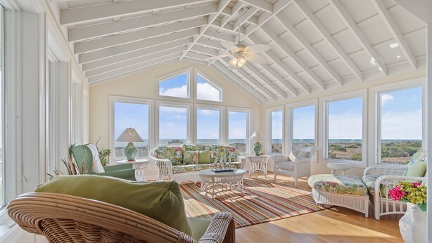 A spacious room with a high ceiling, large windows, wicker furniture, and a view of the landscape. - "Goins My Way" 6 Bedrooms, 6.5 Bathrooms, Private Pool and Elevator (Holden Beach)