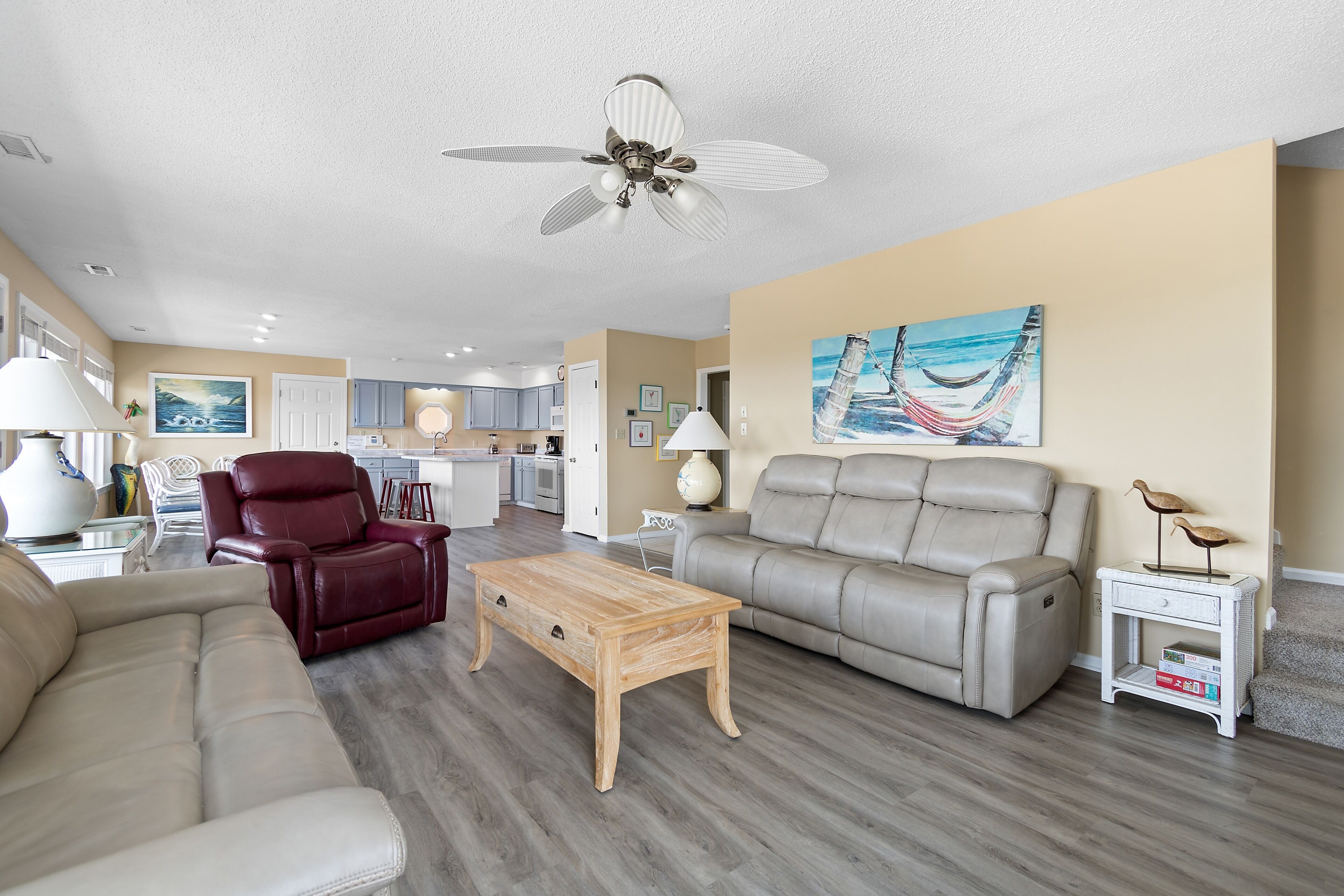 A living room with a beige sofa, a red leather chair, a wooden coffee table, a beige sofa, a ceiling fan, and a painting of a boat on the wall.