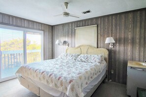 Un dormitorio con cama, ventilador de techo, ventana con persianas, mesita de noche y baño con lavamanos.