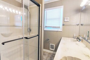 Un baño con cabina de ducha de vidrio, tocador con lavamanos y ventana con persianas.