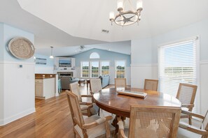 Un'ampia zona pranzo con un grande tavolo di legno e sedie in vimini, una cucina sullo sfondo e una ventola a soffitto.