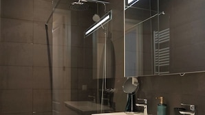 A modern bathroom with a glass shower enclosure, a white vanity with a mirror, and a dark tiled wall. - Cozy apartment canal view (Amsterdam)
