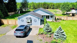 A single-story house with a car parked in the driveway, surrounded by a fenced yard and a forested area. - Large 6BR with Game Room, Perfect for Large Families & Groups, Forks PNW Retreat (Forks)