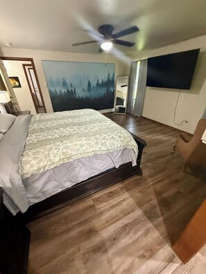 Une chambre à coucher avec un lit, un ventilateur de plafond, un tableau, un miroir et un téléviseur fixé au mur.