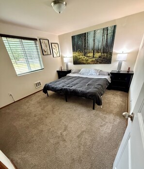 Un dormitorio con cama, dos mesitas de noche, un cuadro grande con tema de bosque, una ventana con persianas y una puerta.