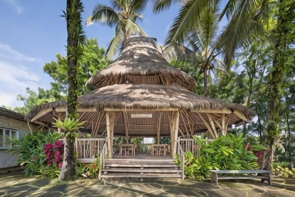 Un pavillon à toit de chaume, doté d’un banc en bois et d’une petite table, entouré d’une végétation luxuriante et de palmiers.