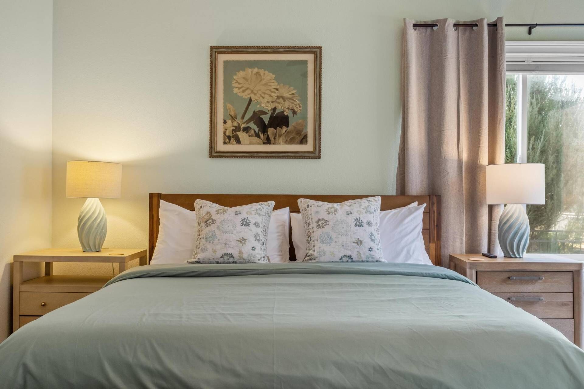 Un lit bien fait, recouvert d’une couverture verte, deux lampes de chevet, un tableau floral encadré accroché au mur et une fenêtre avec des rideaux.