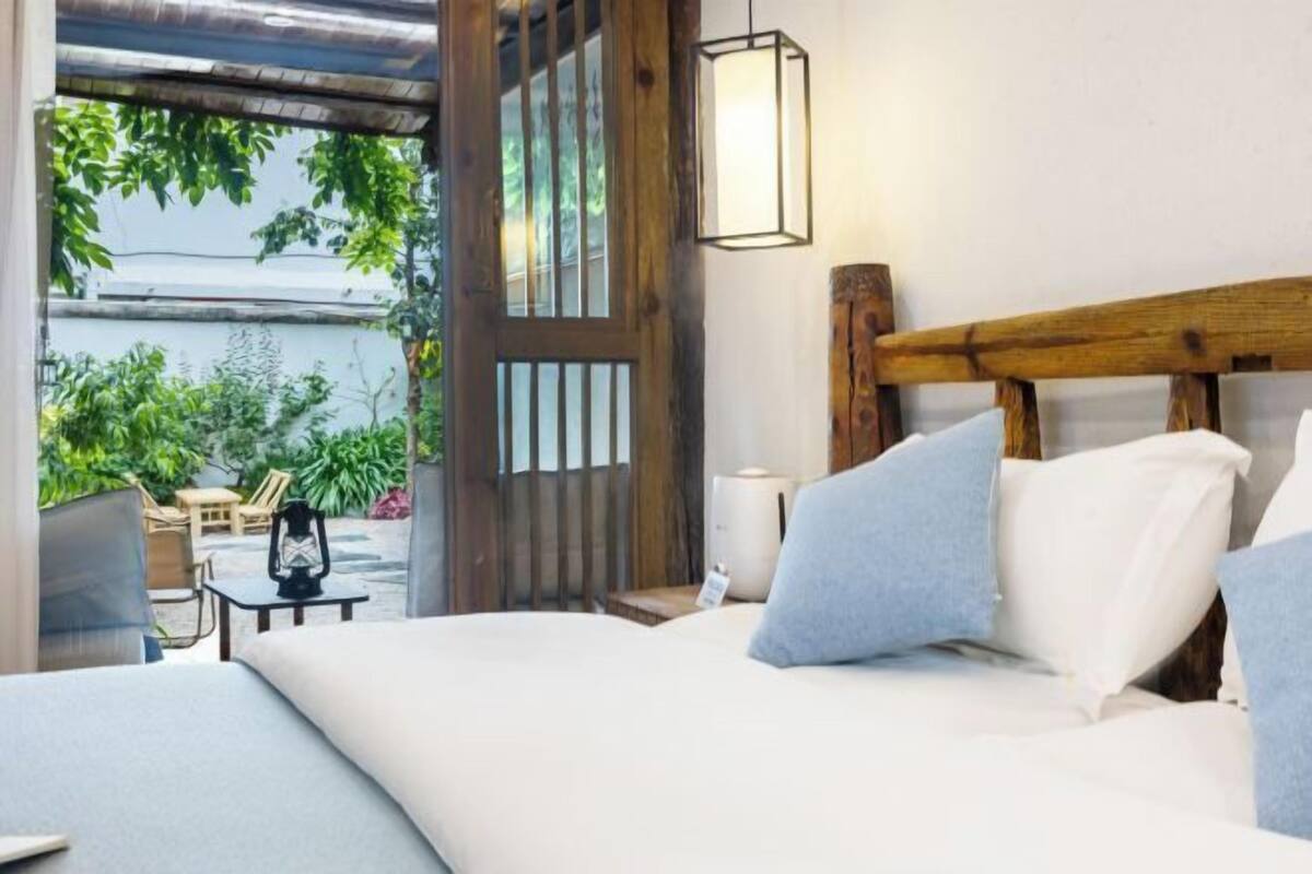 A bedroom with a wooden headboard, a bedside table with a lamp, and a view of an outdoor patio with plants.
