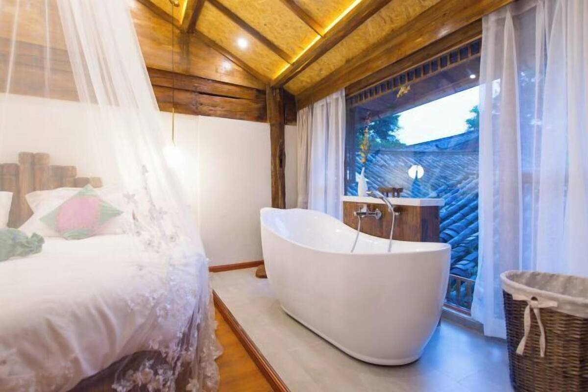 A bedroom with a bed, a white bathtub, and a window with curtains.