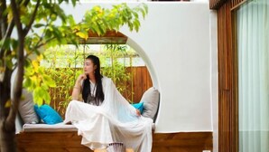 A person sitting on a wooden bench with cushions, under a white structure with a curved top, surrounded by greenery. - ‌The Secret Garden of Cats & Wolf's Den Barista Courtyard Cafe & Inn (Wuyi Street) (Lijiang)
