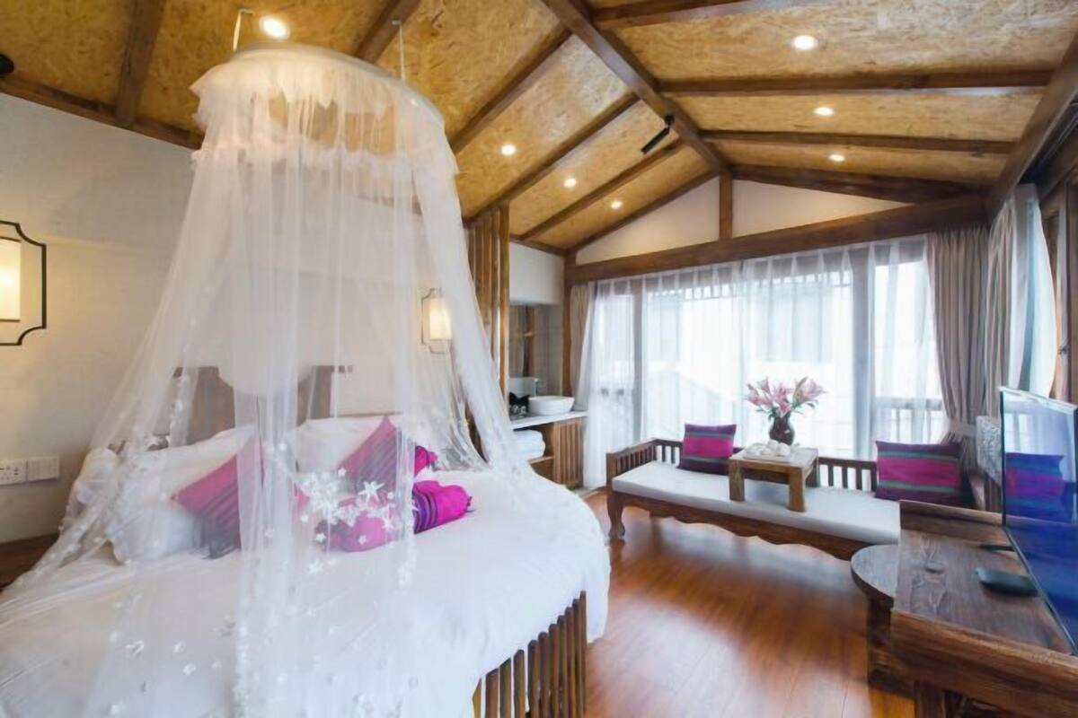 A room with a bed under a canopy, a wooden ceiling, a dining area with a table and chairs, and a large window with curtains.